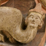 Eine mittelalterliche Steinskulptur eines Kamels mit menschenähnlichem Gesicht und Hut, mit gebogenem Hals und stilisierten Details, wahrscheinlich Teil einer architektonischen Dekoration. (Text mit KI erstellt)