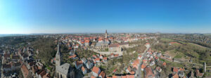 Ein Panoramablick auf eine historische europäische Stadt mit roten Dächern, Kirchtürmen, Steinmauern, Grünflächen und einem weiten blauen Himmel bei klarem Tageslicht. (Text mit KI erstellt)