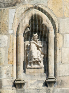Eine Steinstatue einer sitzenden Figur, möglicherweise einer Königin oder einer Heiligen, steht in einer gewölbten Nische auf einer Steinmauer und ist durch ein Netz geschützt. Die Wand besteht aus verschiedenfarbigen Steinblöcken. (Text mit KI erstellt)