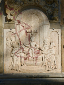 Steinrelief mit einer zentralen Figur auf einem Thron, die von fünf Personen umgeben ist. Das Kunstwerk ist mit roter Farbe bespritzt, was auf Vandalismus schließen lässt. Der Hintergrund ist mit architektonischen Details verziert. (Text mit KI erstellt)