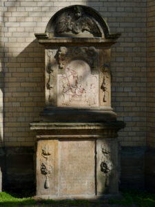 Ein verziertes Steindenkmal mit eingravierten Figuren und Text vor einer hellen Backsteinmauer. Das Relief stellt eine religiöse Szene mit mehreren Personen dar, und die Struktur ist mit dekorativen Schnitzereien und Engelsfiguren versehen. (Text mit KI erstellt)
