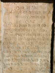Alte, verwitterte Steintafel mit eingraviertem deutschen Text, der teilweise verblasst und schwer lesbar ist, mit einem Gebet an Maria und religiösen Hinweisen. Die Plakette weist Alters- und Erosionsspuren auf. (Text mit KI erstellt)