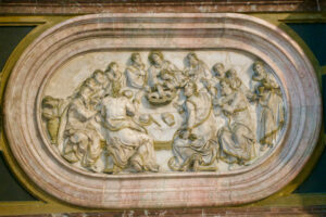 Ein Marmorrelief mit der Darstellung des letzten Abendmahls, bei dem Jesus und zwölf Jünger an einem Tisch sitzen und gemeinsam essen. Das Kunstwerk hat eine ovale Form und ist kunstvoll gerahmt. (Text mit KI erstellt)