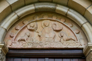 Ein Steinrelief über einem gewölbten Portal zeigt eine sitzende Figur mit Heiligenschein in der Mitte, flankiert von Tierfiguren und umgeben von kreisförmigen Motiven, die möglicherweise eine religiöse oder historische Szene darstellen. (Text mit KI erstellt)