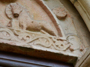 Steinrelief mit der Darstellung eines Löwen, der über kunstvollem Blattwerk steht und hinter seinem Kopf einen Heiligenschein trägt, daneben eine menschliche Figur mit einem Helm. Die Schnitzerei ist in einen gewölbten architektonischen Rahmen eingefasst. (Text mit KI erstellt)