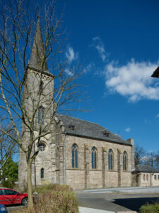 Eine steinerne Kirche mit einem hohen Kirchturm und gewölbten Fenstern steht unter einem strahlend blauen Himmel mit vereinzelten Wolken. Im Vordergrund sind ein blattloser Baum und ein rotes Auto zu sehen. (Text mit KI erstellt)