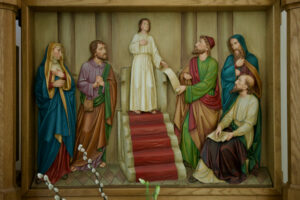 Ein geschnitztes und bemaltes Holzrelief zeigt einen jungen Jesus, der stehend zu einer Gruppe von Erwachsenen in Gewändern spricht; sie hören aufmerksam zu, einige sitzend, andere stehend, in der Mitte eine Treppe mit rotem Teppich. (Text mit KI erstellt)