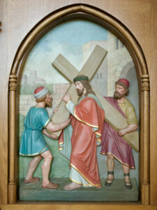 Das Relief zeigt Jesus, der ein Kreuz trägt, ein rotes Gewand und eine Dornenkrone trägt und von einem Mann in Blau unterstützt wird, während ein anderer Mann in Gelb hinter ihnen geht. Die Szene wird von einer gewölbten Holzleiste umrahmt. (Text mit KI erstellt)