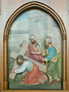 Eine Reliefskulptur zeigt Jesus kniend und ein Holzkreuz tragend, unterstützt von zwei Männern, alle in Gewänder gekleidet, vor einem gewölbten Hintergrund mit steinernen Gebäuden und Grünpflanzen. (Text mit KI erstellt)