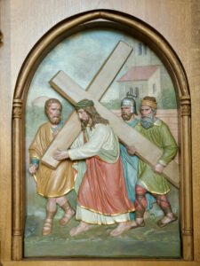 Das Relief zeigt Jesus, der ein großes Holzkreuz trägt, begleitet von drei Männern, von denen zwei ihm helfen. Sie sind in Gewänder gekleidet und im Hintergrund ist ein Gebäude mit Bäumen zu sehen. (Text mit KI erstellt)