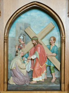 Eine Reliefskulptur zeigt Jesus, der ein Holzkreuz trägt, und eine Frau, die kniet und seine Hand berührt. Zwei Männer, einer in Rüstung, stehen hinter ihnen. Die Szene wird von einem Holzbogen eingerahmt. (Text mit KI erstellt)