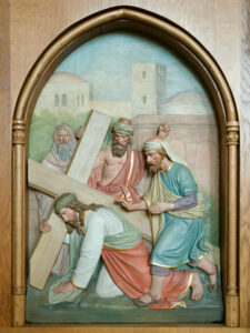 Eine Reliefskulptur zeigt Jesus, der ein Holzkreuz trägt und von drei Männern umgeben ist, von denen einer ihm hilft und die beiden anderen ihn beobachten, vor dem Hintergrund einer antiken Stadt in einem gewölbten Holzrahmen. (Text mit KI erstellt)