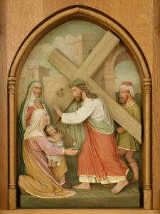 Eine Reliefskulptur zeigt Jesus, der ein Kreuz trägt und auf seinem Weg Frauen und einem Kind begegnet. Jesus reicht ihnen die Hand, während zwei Frauen, von denen eine ein Kind hält, vor ihm knien. Dahinter stehen zwei Männer in biblischen Gewändern vor einem gewölbten Rahmen. (Text mit KI erstellt)