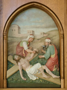 Ein Relief zeigt Jesus, der an einem Holzkreuz liegt, während ein Mann seine Hand festnagelt und ein anderer assistiert, vor einer hügeligen Landschaft mit Gebäuden im Hintergrund, eingerahmt von einem Holzbogen. (Text mit KI erstellt)