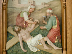 Eine Reliefskulptur zeigt Jesus am Kreuz, während sich ein Mann darauf vorbereitet, seine Hand festzunageln, während eine Frau in der Nähe steht und den Arm Jesu hält. Die Szene findet im Freien statt, mit Gebäuden im Hintergrund und eingerahmt von Holz. (Text mit KI erstellt)