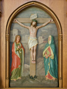 Eine Reliefskulptur der Kreuzigung Jesu mit Jesus am Kreuz in der Mitte, flankiert von zwei Figuren in Gewändern auf beiden Seiten, eingefasst in einen hölzernen Bogenrahmen. (Text mit KI erstellt)