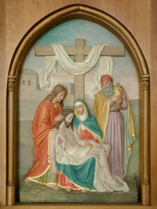 Eine Reliefskulptur zeigt die Pietà-Szene mit dem von Maria gehaltenen Körper Jesu, umgeben von zwei weiteren Figuren, vor einem mit einem Tuch drapierten Holzkreuz. Die Szene wird von einer gewölbten Holzleiste eingerahmt. (Text mit KI erstellt)