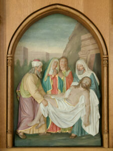 Eine Reliefskulptur zeigt vier Personen, die den in ein weißes Tuch gehüllten Leichnam von Jesus Christus in das Grab legen. Die Trauernden tragen Gewänder und stehen vor einem steinernen Eingang und einem grünen Berghang. (Text mit KI erstellt)