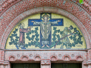 Ein Mosaik in einem gemauerten Bogen zeigt Jesus mit ausgestreckten Armen zwischen zwei Figuren: links eine, die einen Kelch anbietet, und rechts eine, die Weinreben pflegt, wobei grüne Ranken ein verziertes Muster hinter ihnen bilden. (Text mit KI erstellt)
