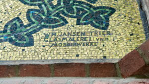 Ein Mosaik mit einem grünen und blauen geometrischen Muster auf einem Hintergrund aus kleinen gelben Fliesen. Der Text lautet: W.H. Jansen Trier Glas Malerei u[nd] Mosaikwerke. Der Rand ist mit roten Ziegeln eingefasst. (Text mit KI erstellt)