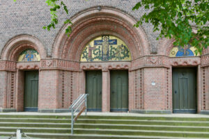 Drei gewölbte Portale eines Backsteingebäudes, jedes mit einer grünen Tür und einem Mosaik darüber. Das zentrale Mosaik stellt eine Figur am Kreuz dar, die seitlichen Mosaike zeigen weitere Figuren. Eine Treppe und ein Metallgeländer führen zum Eingang hinauf. (Text mit KI erstellt)