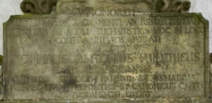 Eine verwitterte Steintafel mit lateinischen Inschriften, deren Text teilweise abgenutzt und schwer lesbar ist, erinnert an ein Ereignis oder eine Person in der Sakramentskapelle Fronleichnam in Büren. Die Gravur weist am oberen Rand dekorative Elemente auf. (Text mit KI erstellt)