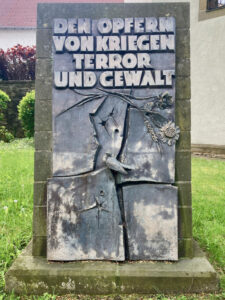Eine steinerne Gedenktafel mit der erhabenen deutschen Aufschrift Den Opfern von Kriegen, Terror und Gewalt, umgeben von Gras, Blumen und einem gemalten Vogel, ist den Opfern von Krieg, Terror und Gewalt gewidmet. (Text mit KI erstellt)