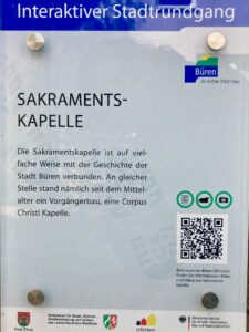 Ein Informationsschild in deutscher Sprache über die Sakramentskapelle in Büren, mit einer blauen Kopfzeile, dem Stadtlogo und einem Text, der die Geschichte der Kapelle beschreibt. Am unteren Rand befinden sich mehrere Logos und ein QR-Code für weitere Informationen. (Text mit KI erstellt)