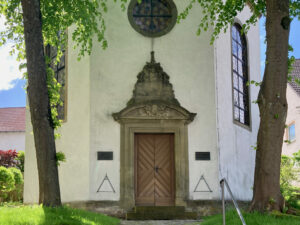 Ein weißes Kirchengebäude mit einem dekorativen Steineingang, hölzernen Doppeltüren und gewölbten Buntglasfenstern, eingerahmt von zwei großen, belaubten Bäumen auf beiden Seiten. Das Sonnenlicht fällt durch die grünen Blätter. (Text mit KI erstellt)