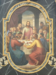 Ein klassisches Gemälde zeigt Jesus in der Mitte eines langen Tisches, umgeben von seinen Jüngern, die in einem kunstvollen, gewölbten Rahmen mit marmorähnlicher Umrandung und dekorativen Akzenten ein Mahl halten. (Text mit KI erstellt)