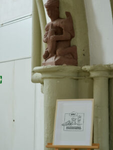Eine gerahmte Illustration auf einer Staffelei steht unter einer roten Steinstatue einer sitzenden Figur, die einen Gegenstand hält, montiert auf einer Säule in einem Innenraum mit hellen Wänden. (Text mit KI erstellt)
