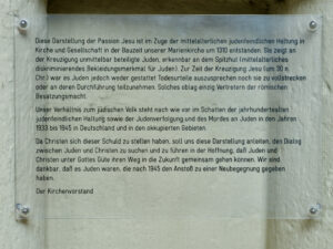 Eine Tafel mit deutschem Text, der den historischen Kontext einer Passionsdarstellung erklärt, Antisemitismus anspricht und das Gedenken an den Holocaust fördert, ist an einer Wand mit sichtbaren Schrauben an jeder Ecke angebracht. (Text mit KI erstellt)
