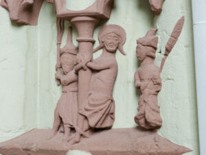 Ein Steinrelief, das eine an eine Säule gefesselte Figur mit Heiligenschein darstellt, die von zwei anderen Figuren flankiert wird; eine scheint eine Peitsche zu halten, was auf eine Szene der Geißelung oder Bestrafung hindeutet. Die Skulptur ist verwittert und steht auf einer strukturierten Wand. (Text mit KI erstellt)