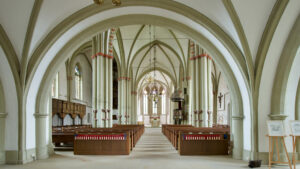Innenansicht einer Kirche mit hohen Gewölbedecken, Reihen von Holzbänken, Kronleuchtern, Säulen und religiösen Symbolen an den Wänden, die zum Altar am anderen Ende führen. (Text mit KI erstellt)