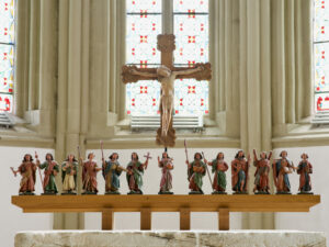 Ein Holzaltar mit 13 farbenfrohen Statuen von Figuren, die verschiedene Gegenstände halten, steht unter einem großen Kruzifix mit Jesus vor hohen Buntglasfenstern in einer Kirche. (Text mit KI erstellt)