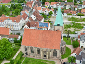 Luftaufnahme einer großen Steinkirche mit rotem Ziegeldach und grünem Kirchturm, umgeben von Bäumen und Wohngebäuden mit ähnlichen roten Dächern in einer Kleinstadt. Auf einem nahe gelegenen Gebäude ist ein Sonnenkollektor zu sehen. (Text mit KI erstellt)