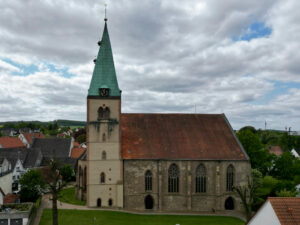 Eine große Steinkirche mit einem hohen Glockenturm mit grüner Spitze steht zwischen Häusern unter einem bewölkten Himmel. Die Kirche hat Rundbogenfenster und ein braunes Ziegeldach und ist von grünen Rasenflächen und Bäumen umgeben. (Text mit KI erstellt)