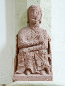 Eine verwitterte Steinskulptur einer sitzenden Figur mit gekreuzten Armen und Beinen, die in einer einfachen, stilisierten Art und Weise vor einer hellen Wand gemeißelt ist. (Text mit KI erstellt)