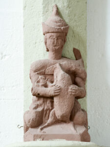 Eine Statue aus rötlichem Stein mit einer sitzenden Figur, die einen spitzen Hut trägt und einen großen Fisch auf der Brust hält, steht vor einer hellen Wand. (Text mit KI erstellt)