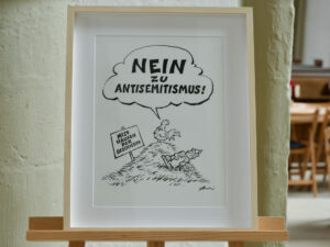 Eine gerahmte Karikatur zeigt einen Hahn, der in einer Sprechblase NEIN ZU ANTISEMITISMUS! (NEIN ZUM ANTISEMITISMUS!) in einer Sprechblase. Auf einem Schild daneben steht MIST HÄUFEN DER GESCHICHTE. (Text mit KI erstellt)