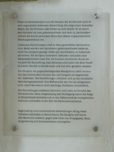 Eine Tafel mit deutschem Text erklärt die antisemitische Symbolik einer mittelalterlichen Sandsteinskulptur an einer Kirche, die Juden mit Schweinen abbildet. Der Text kritisiert diese Geschichte und ruft dazu auf, Antisemitismus und solche Bilder heute abzulehnen. (Text mit KI erstellt)