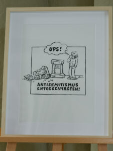Eine gerahmte Cartoon-Zeichnung zeigt eine Person, die auf einen vandalisierten Grabstein blickt und UPS! sagt. Der Grabstein und die umliegenden Trümmer sind mit Graffiti beschmiert. Der Text darunter lautet: ANTISEMITISMUS ENTGEGENTRETEN! (Steht auf gegen Antisemitismus!). (Text mit KI erstellt)