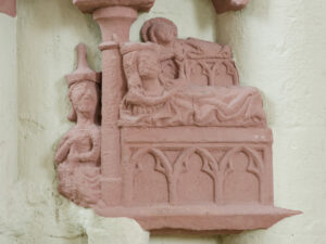 Ein rötliches Steinrelief zeigt zwei liegende Figuren auf einem Bett im gotischen Stil, daneben eine weitere sitzende Figur vor einem hellen Hintergrund. (Text mit KI erstellt)
