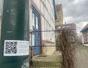Ein QR-Code-Aufkleber an einem grünen Pfahl vor einem Fachwerkhaus mit blauen Türen und Fenstern, an einem bewölkten Tag in einer historisch anmutenden Straße. (Text mit KI erstellt)