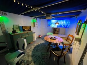 Ein gemütlicher, maritimer Escape-Room in Warburg mit Lichterketten, einem runden Holztisch mit verschiedenen Stühlen, einem Vintage-Radio auf einem grünen Schrank, einem Teppich mit Meeresmotiven und einer blauen Meeresszene, die auf die Rückwand projiziert wird. (Text mit KI erstellt)