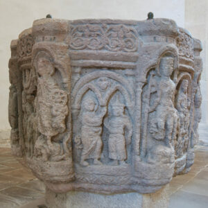 Ein achteckiges Taufbecken aus Stein mit kunstvollen Schnitzereien von menschlichen Figuren und Bögen auf jeder Platte in einem Innenraum mit Steinfußboden und einer weißen Wand im Hintergrund. (Text mit KI erstellt)