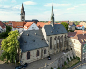 Luftaufnahme einer historischen Steinkirche mit steilem Schieferdach und hohen Rundbogenfenstern, eingebettet zwischen rotgedeckten Gebäuden und einem Kirchturm in der malerischen europäischen Stadt Warburg, ada.kreis-höxter. (Text mit KI erstellt)