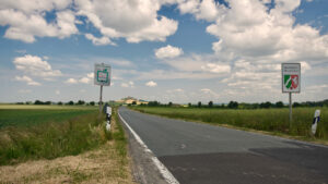 Eine Landstraße schlängelt sich unter teilweise bewölktem Himmel durch grüne Felder. Zwei Schilder auf der rechten Seite markieren die Grenze von Nordrhein-Westfalen, Deutschland. Am Horizont sind in der Ferne landwirtschaftliche Gebäude zu erkennen. (Text mit KI erstellt)