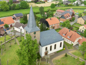 Luftaufnahme eines Dorfes mit einer historischen Kirche mit einem hohen, spitzen Kirchturm, umgeben von Bäumen, Wegen und rotgedeckten Häusern in einer grünen, ländlichen Landschaft. (Text mit KI erstellt)