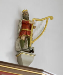 Statue eines bärtigen Königs mit Krone, rotem Gewand und grünen Stiefeln, der auf einem Sockel steht und eine große goldene Harfe vor einem schlichten weißen Hintergrund spielt. (Text mit KI erstellt)